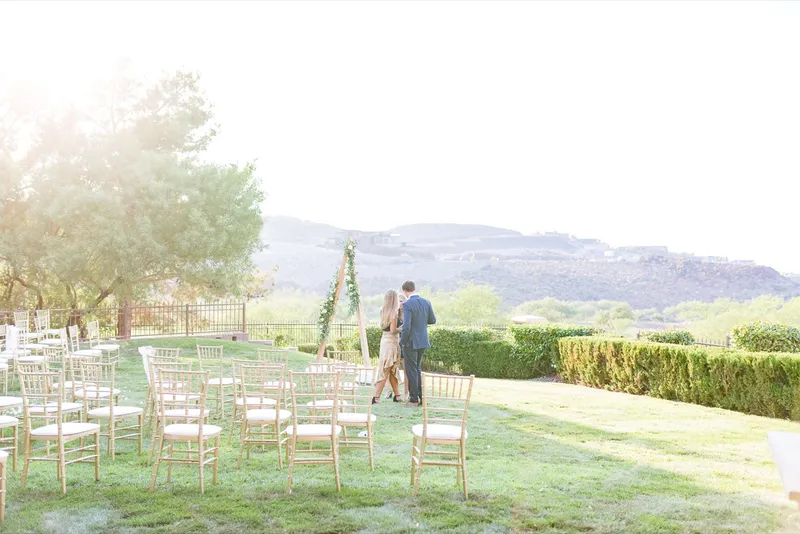 Two guests standing beside the triangle arch at the empty ceremony site, the hills and the valley opening up behind the hedge