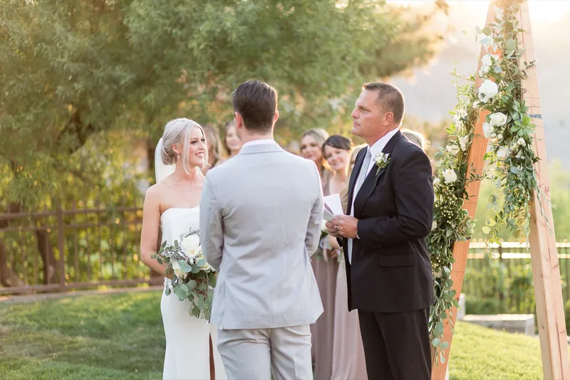 Bride and groom holding hands facing the officiant at the greenery arch, guests seated in front of them