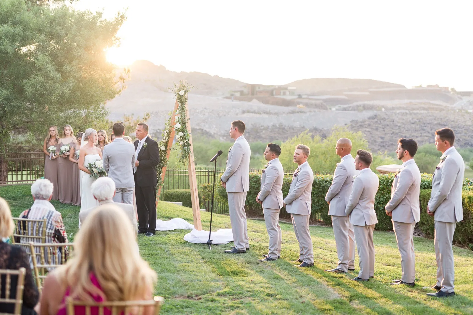 A ceremony on a lawn as the sun drops behind the hills, groomsmen in grey suits lined up along the aisle