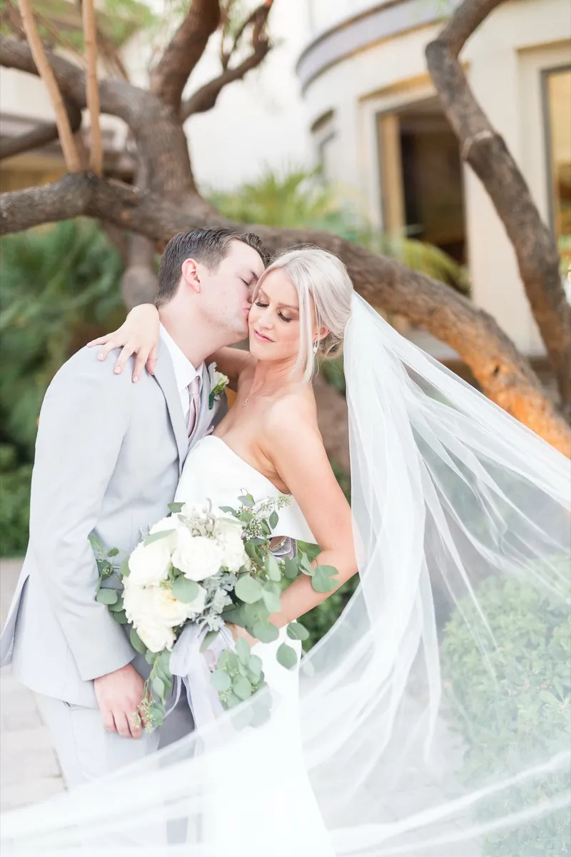 Groom kissing the bride's cheek as her long veil lifts and blows across the frame