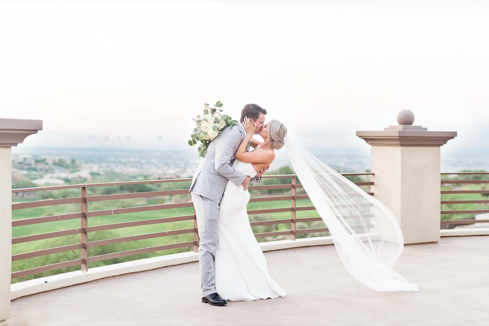 A groom dips his bride for a kiss on a hillside terrace, her veil flying, the valley spread out behind