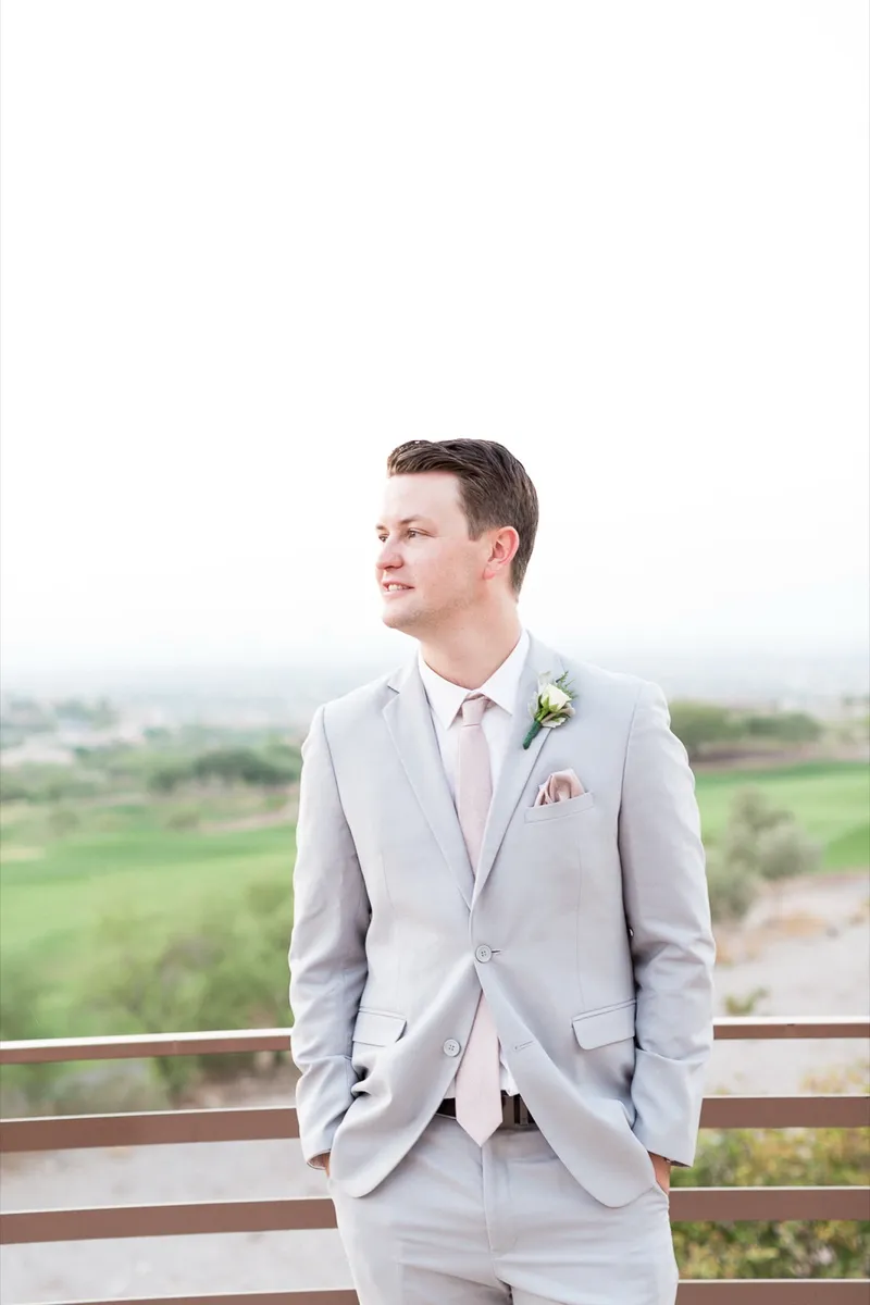 Groom standing at the terrace rail in a light grey suit and blush tie, hands in his pockets