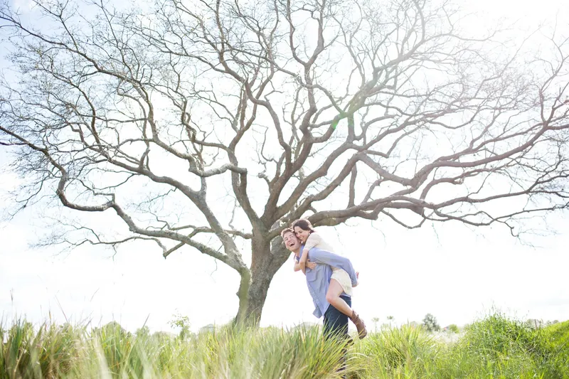Groom carrying his bride to be on his back under a big bare tree