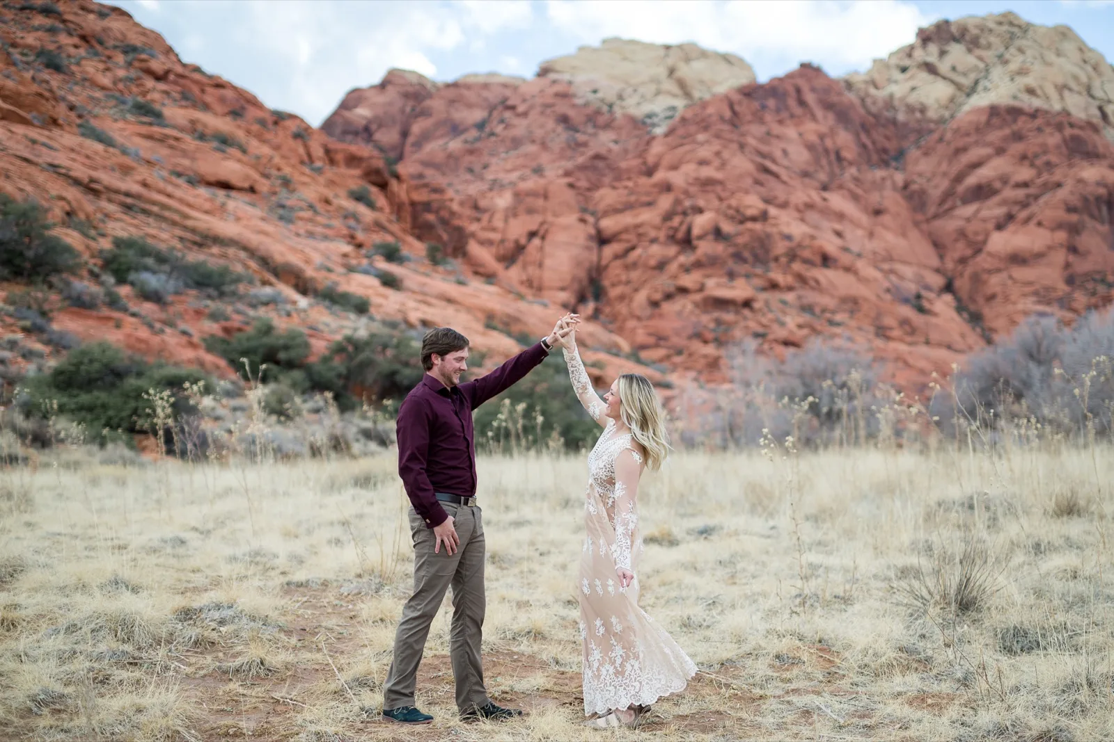 Man in a plum shirt spinning his fiancee in a cream lace dress with the red sandstone cliffs of Red Rock Canyon behind them