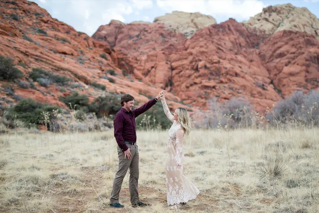 Man in a plum shirt spinning his fiancee in a cream lace dress with the red sandstone cliffs of Red Rock Canyon behind them