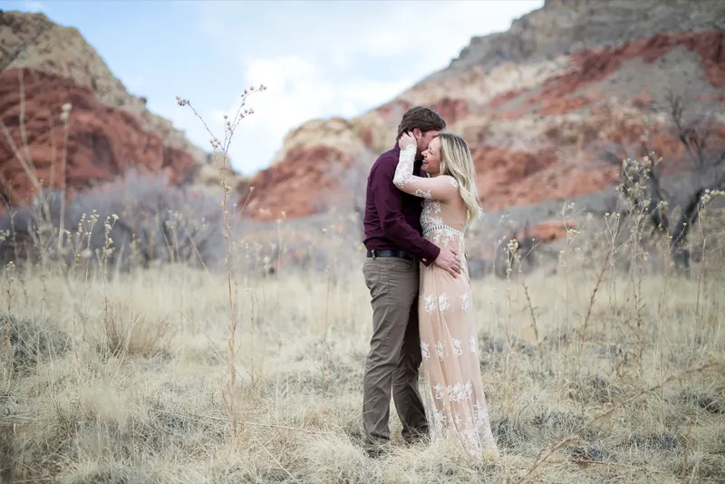 Couple stand forehead to forehead in dry winter grass below the striped red and cream cliffs of Red Rock Canyon