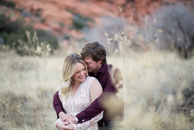 Man kissing his fiancee on the side of her head as she laughs, both of them surrounded by tall golden grass