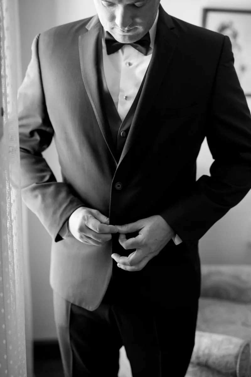 Black and white photo of the groom buttoning his tuxedo jacket in a room with framed pictures on the wall behind him