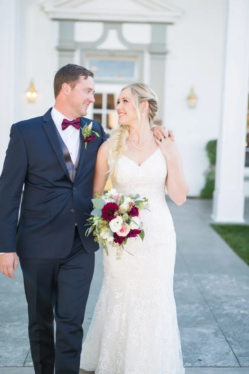 The bride and groom walking arm in arm and looking at each other under the hotel portico, her hand on his shoulder and her bouquet at her side