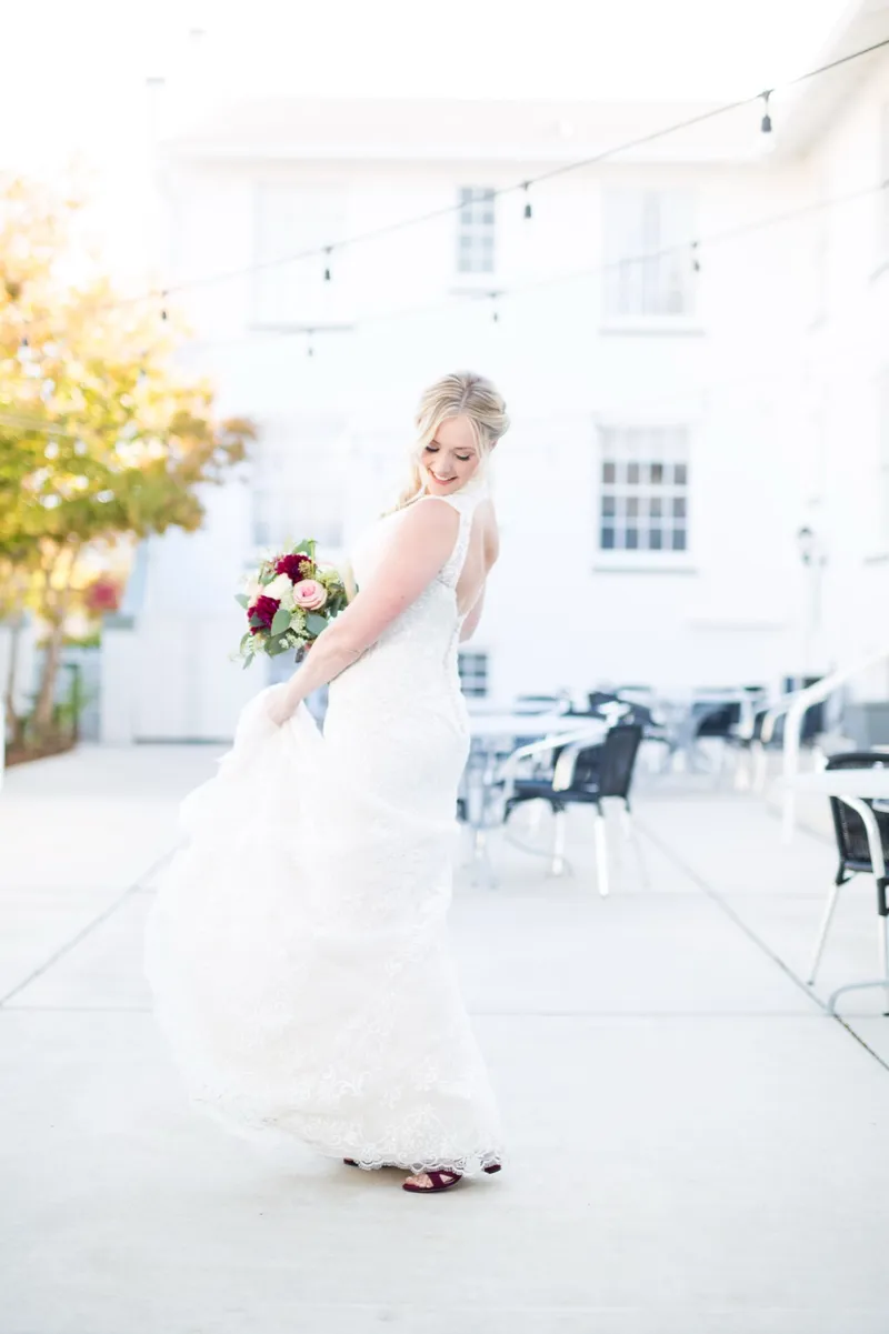 The bride sweeping the lace train of her gown across a concrete patio in burgundy heels, string lights strung overhead and a white two story wall behind her