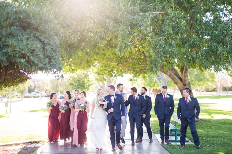 The whole wedding party walking across the park lawn under mature shade trees, bridesmaids in blush and burgundy on one side and groomsmen in navy on the other
