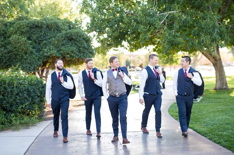 Four groomsmen in navy suits with burgundy ties walking a park path together in dappled afternoon light