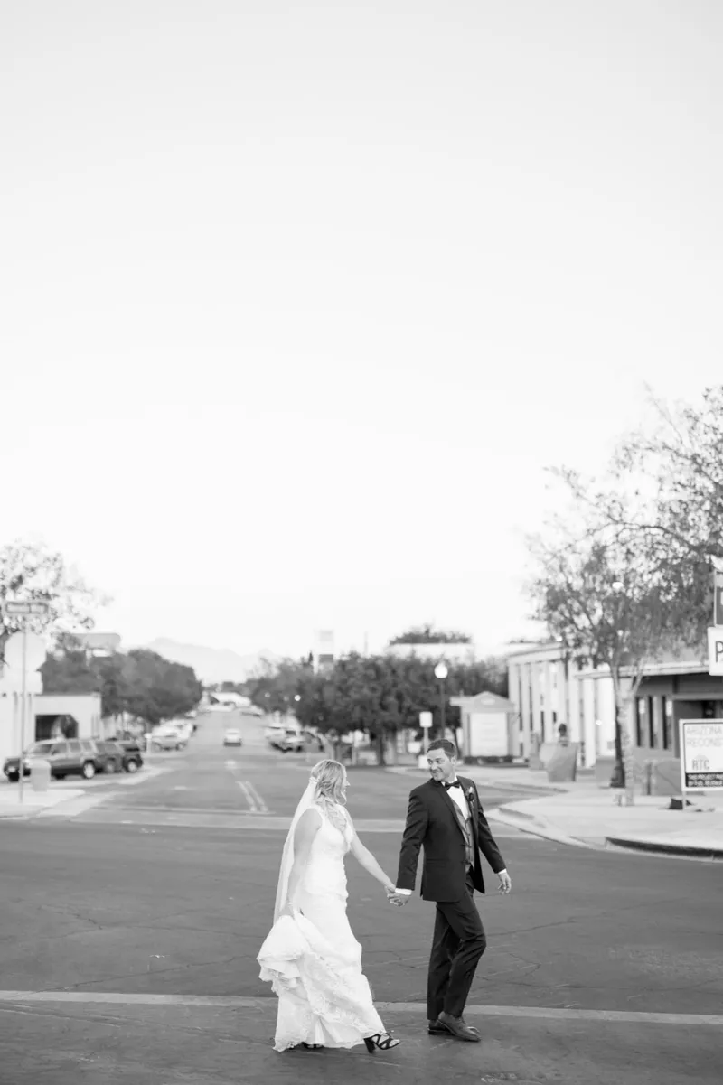 Black and white photo of the bride and groom walking down the middle of a quiet small town street with parked cars and low storefronts on both sides