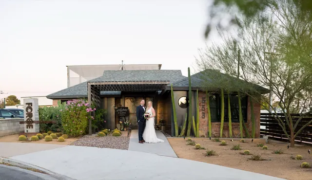 The bride and groom standing together at the venue entrance under the wood pergola at dusk, with barrel cactus, a prickly pear and the metal 553 street numbers on the stone wall beside them