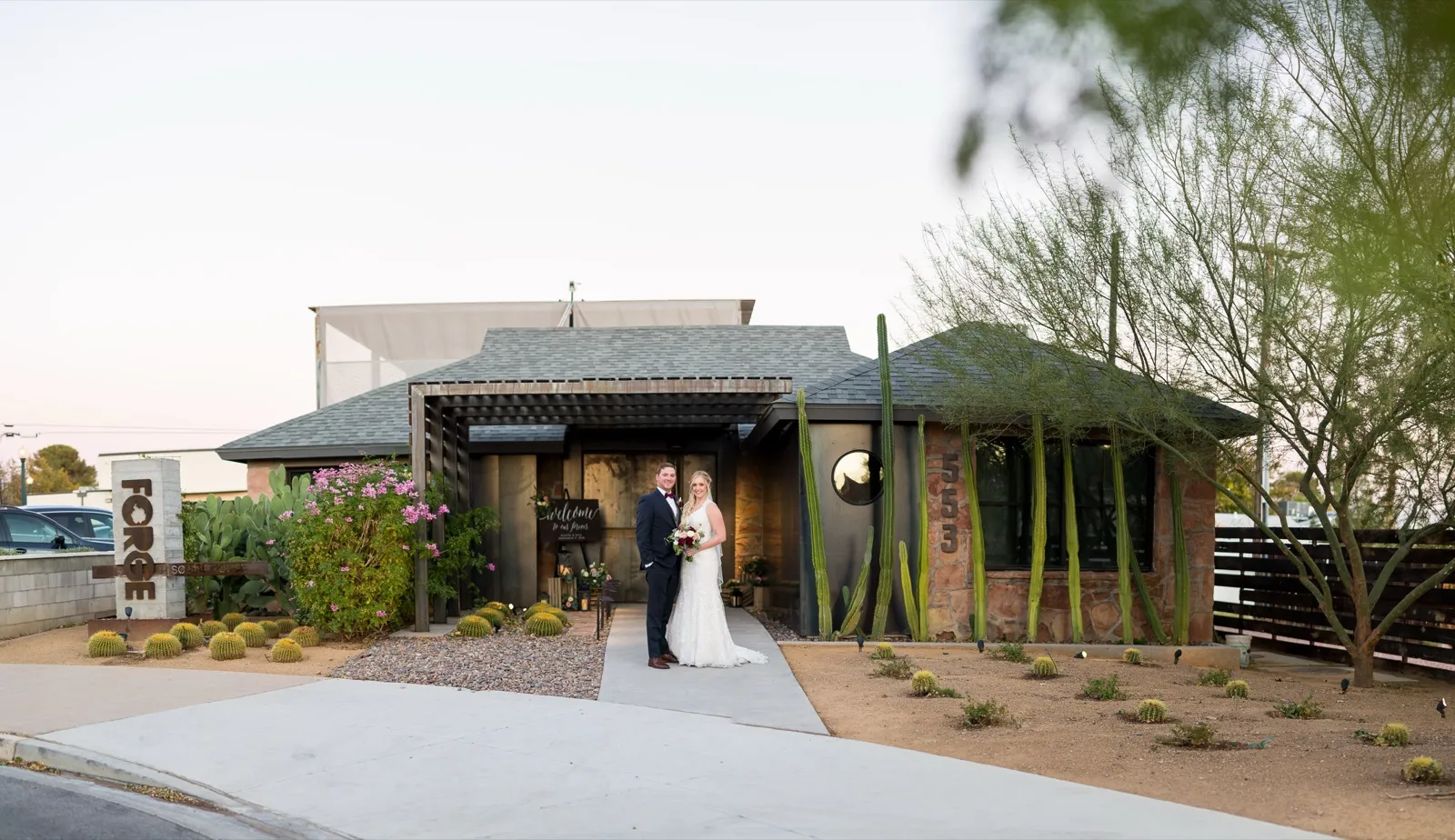 Bride and groom at the lit entrance of a low stone and wood building at dusk, barrel cactus and prickly pear out front