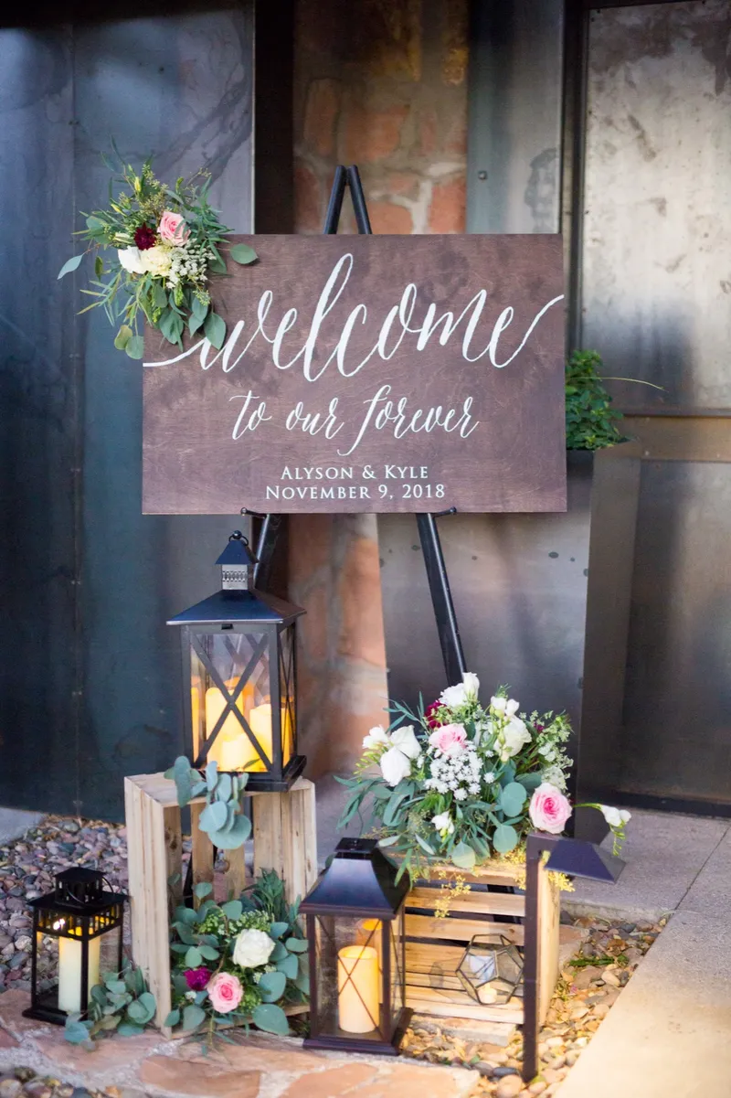 A wood welcome sign lettered in white script standing on an easel against a rusted steel wall, with lanterns and a low arrangement of burgundy and blush flowers stacked at its feet