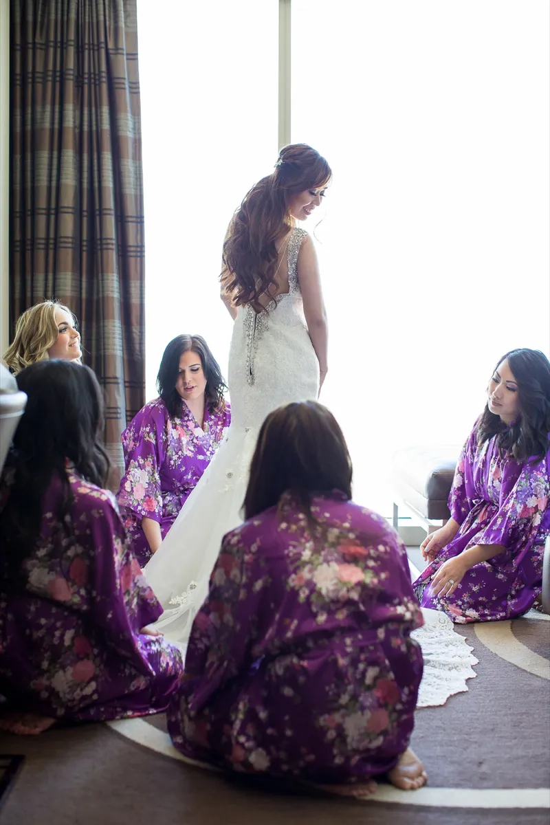 Bride standing in her gown by the window while five bridesmaids in purple floral robes crouch around her and spread the skirt out
