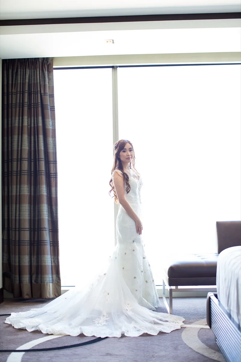 Bride in her gown standing at the floor to ceiling window of the hotel room with her train fanned out across the carpet