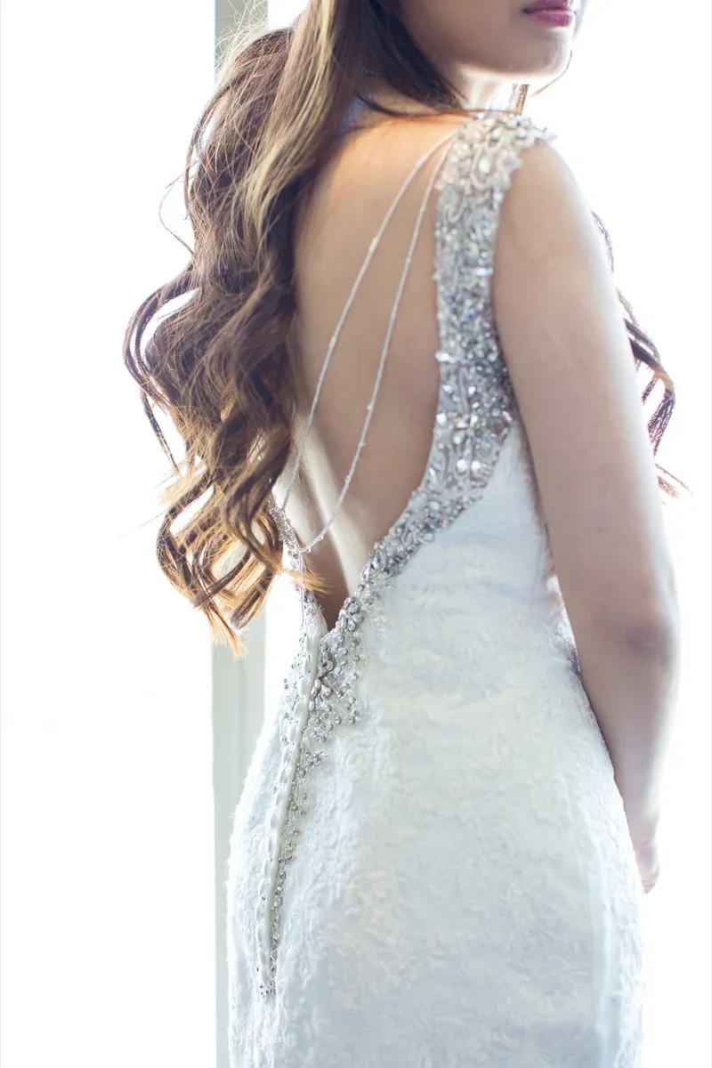 Close view of the open back of the bride's gown with beaded straps crossing her shoulders and her curled hair falling to one side
