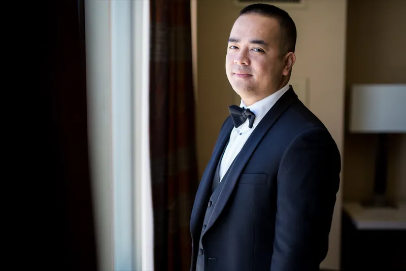 Man in a black tuxedo and bow tie standing beside the hotel room window, turned toward the light