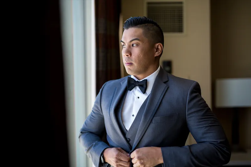 Groom in a charcoal tuxedo buttoning his jacket at the window, looking out