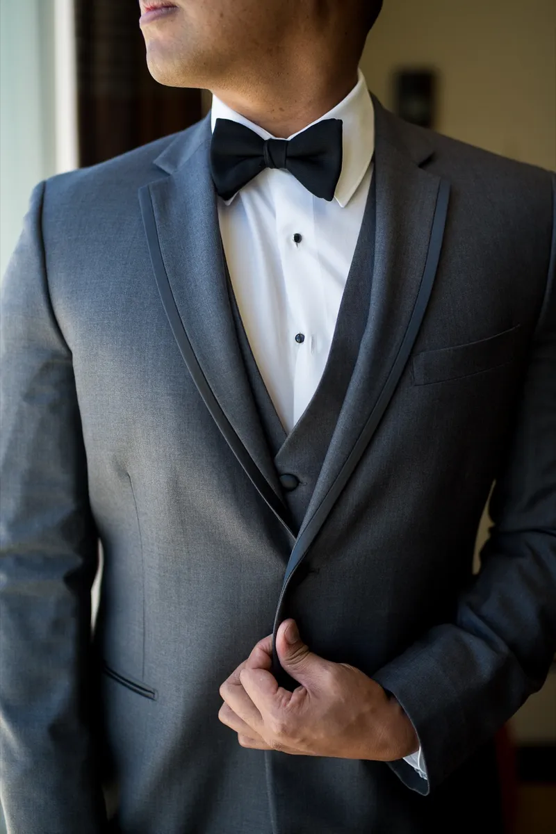 Close crop of the groom's charcoal jacket, matching vest and black bow tie as he fastens a button