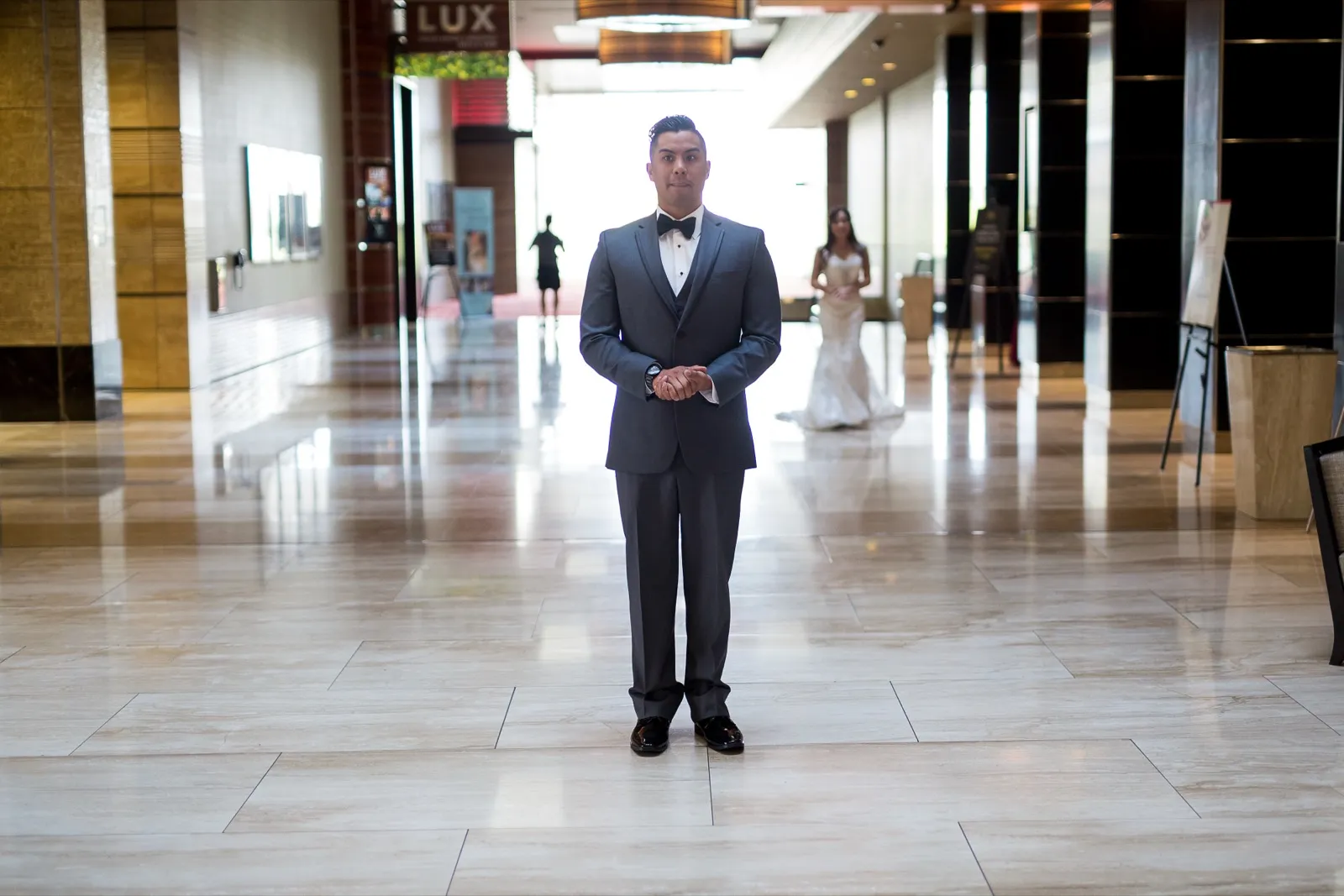 Groom waits alone in an empty resort lobby while his bride walks up behind him