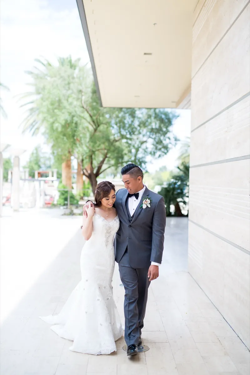 Bride and groom walking together along the shaded entrance drive with palms and a stone wall behind them