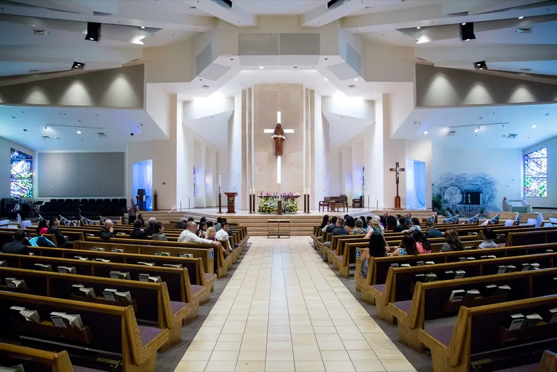 Wide view up the church aisle before the ceremony, guests seated in light wood pews under the tall lit cross above the altar