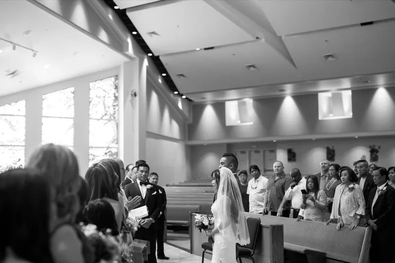 Black and white frame of the bride in her veil reaching the front of the church while the whole congregation stands, daylight coming through the tall side windows