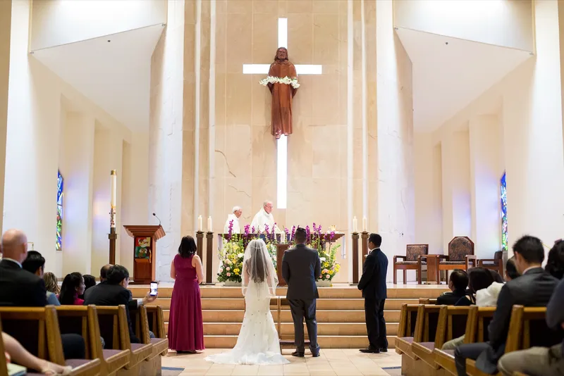 Bride and groom standing at the altar steps with the priest between them and flowers on the step below