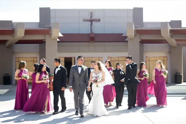 Bride and groom walking across the wide concrete plaza with their wedding party around them, the church entry and the sculpted cross on the front wall behind them