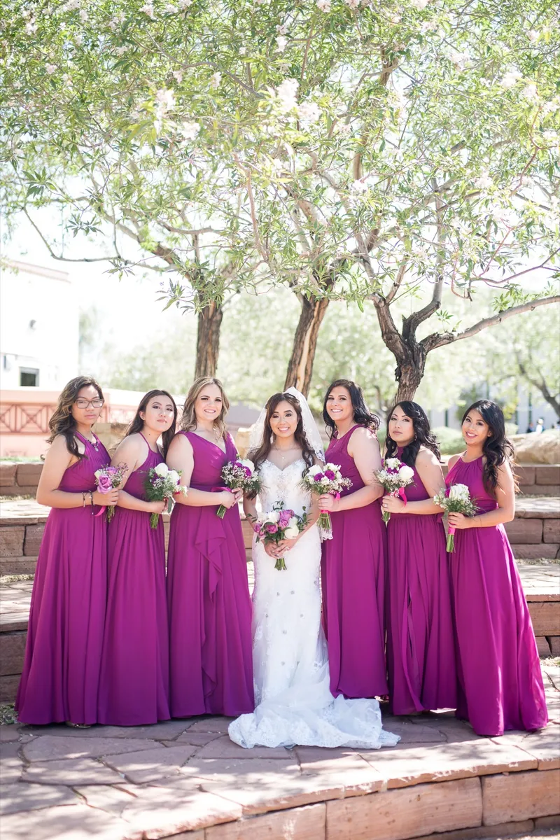 Bride and her bridesmaids in magenta gowns standing together on the stone planter wall under a courtyard tree