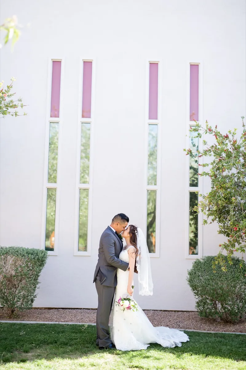 Bride and groom kissing on the grass in front of the tall narrow church windows with rose colored panes at the top