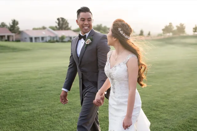 Bride and groom walking hand in hand across the green fairway in low golden light, her hair blowing back and low buildings and trees soft behind them