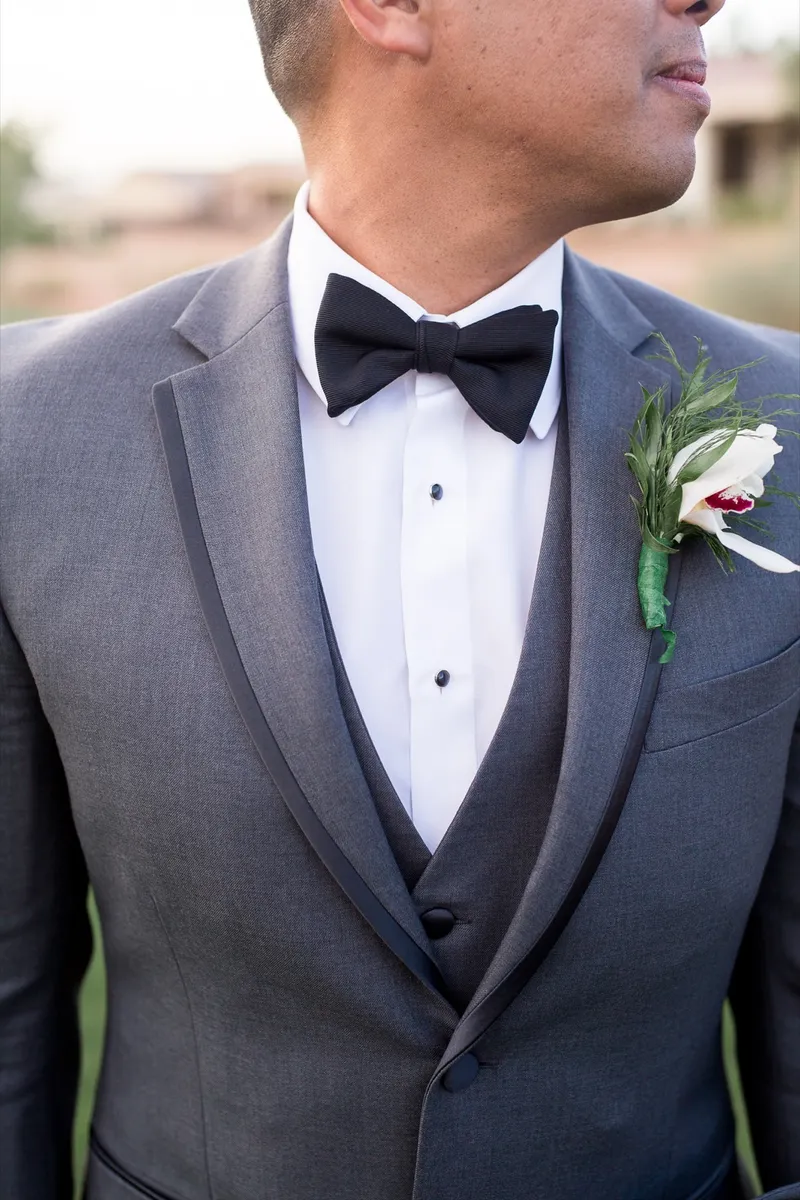 Close view of the groom's grey tuxedo, matching vest and black bow tie with a single white rose boutonniere
