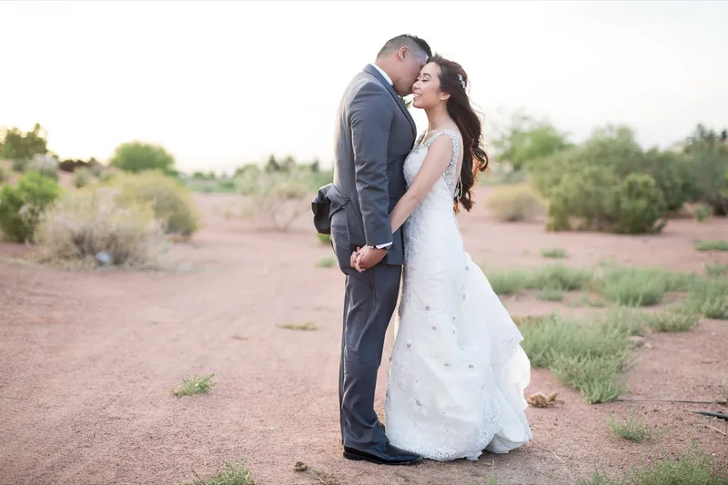 Groom in a grey suit pressing his face to the bride's cheek while she smiles with her eyes closed, the two of them alone on the desert path