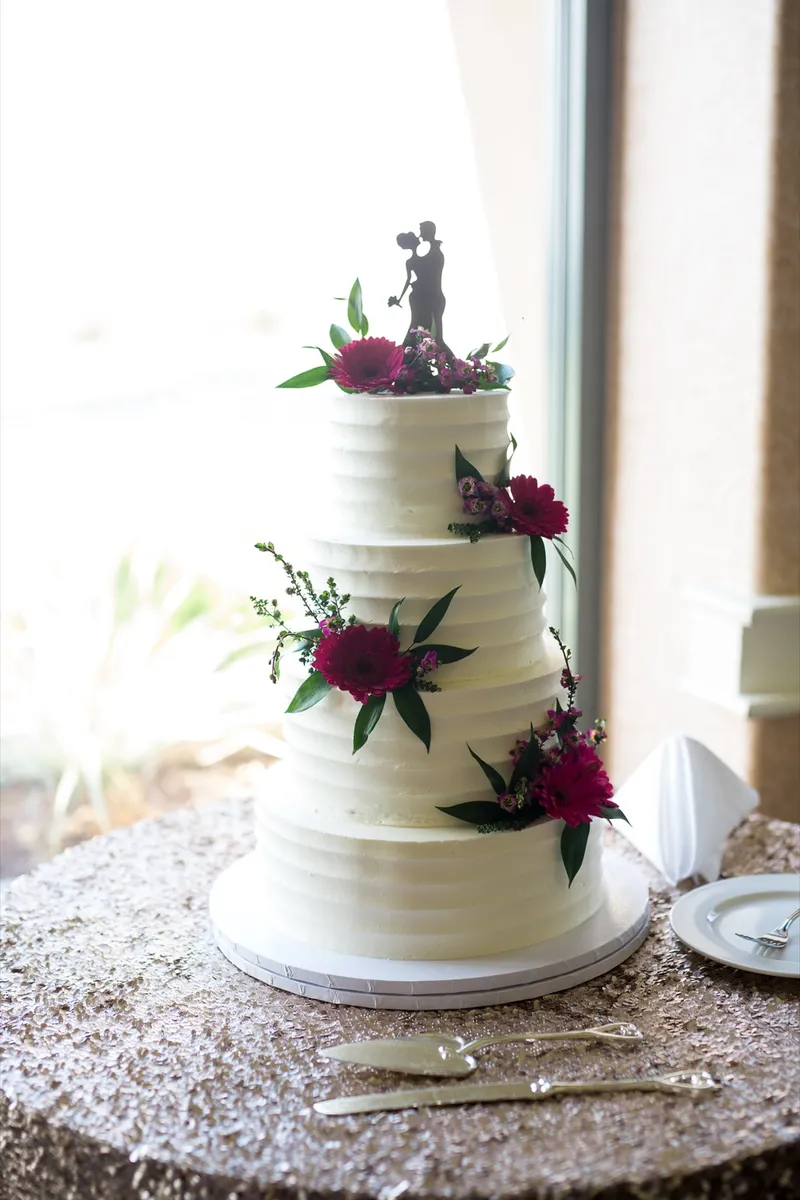 Three tier white wedding cake trimmed with burgundy dahlias and a silhouette topper, standing on a gold sequin cloth beside a tall window