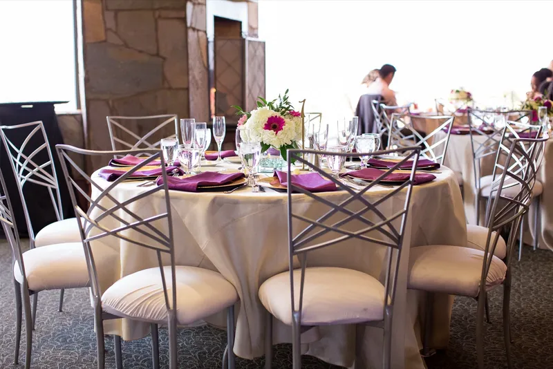 Round reception table in champagne linen with purple napkins on gold chargers and a white and pink centerpiece, silver chairs around it and a stone column behind