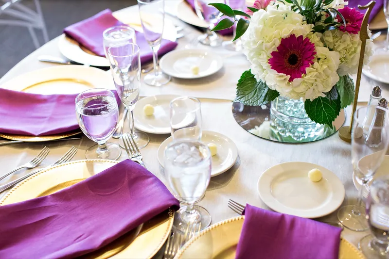 Close view of a place setting with a folded purple napkin on a gold charger, water glasses filled and a white hydrangea and pink gerbera centerpiece behind