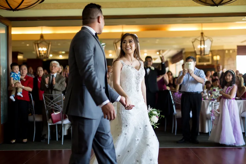 Bride and groom walking onto the dance floor for their entrance while guests stand and applaud, one of them filming on a phone under the lantern lights