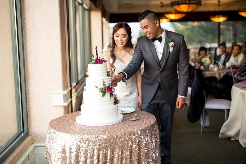 Couple cutting the cake together on its gold sequin table with seated guests and the windows behind them