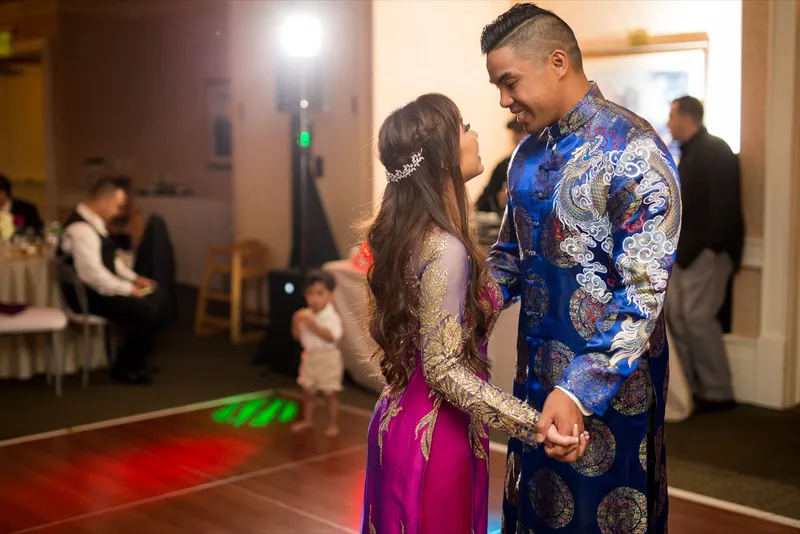 Bride in a pink and gold ao dai and groom in a blue and gold ao dai holding hands on the dance floor under colored lights