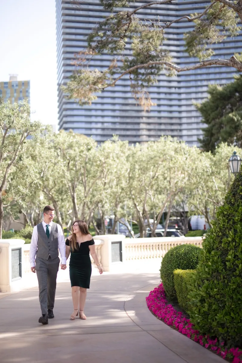 Couple walking a curving path past pink flowers with a blue glass tower rising behind the trees