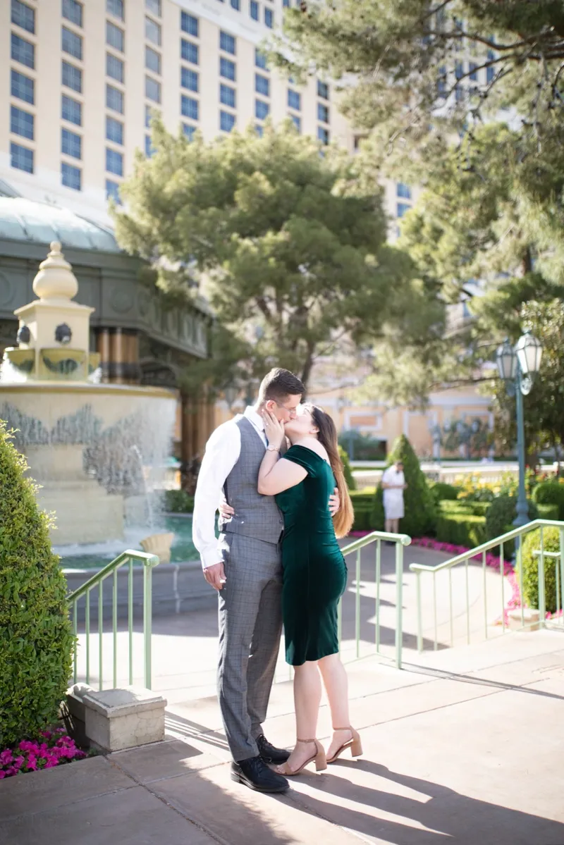 The two of them kissing beside a small fountain with white railings and clipped hedges around them