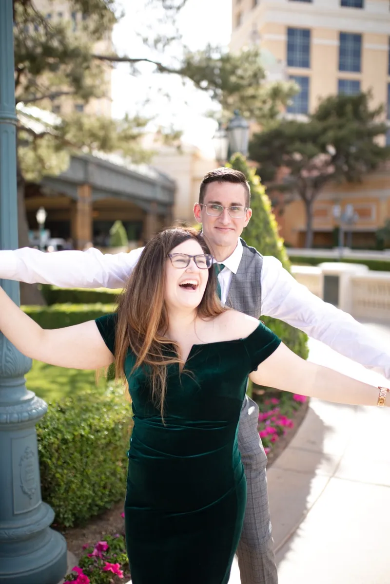 She throws both arms out laughing while he holds her from behind on the garden path