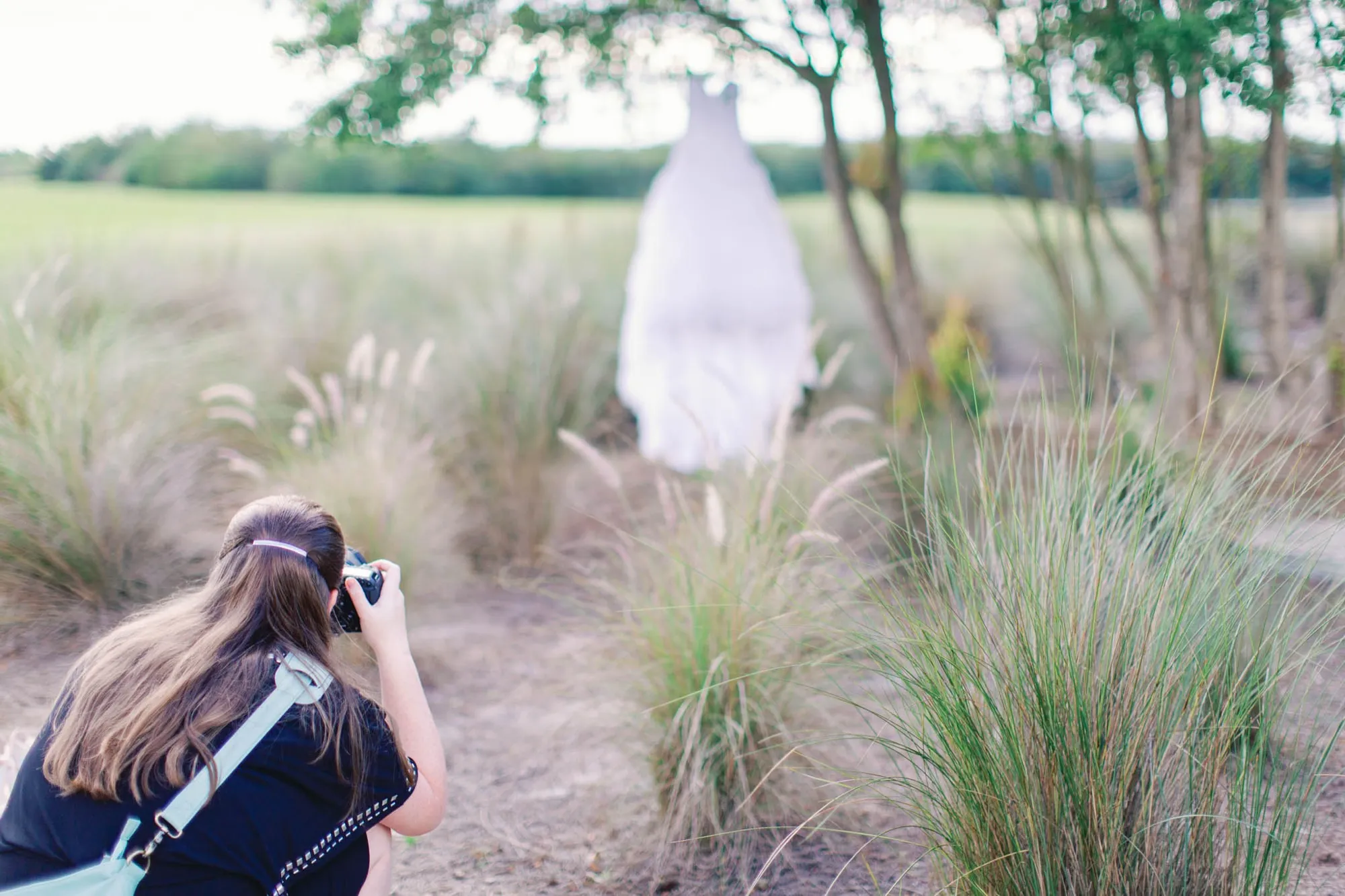 McKenzie crouches low with her camera to photograph a bride standing in tall feathery grasses