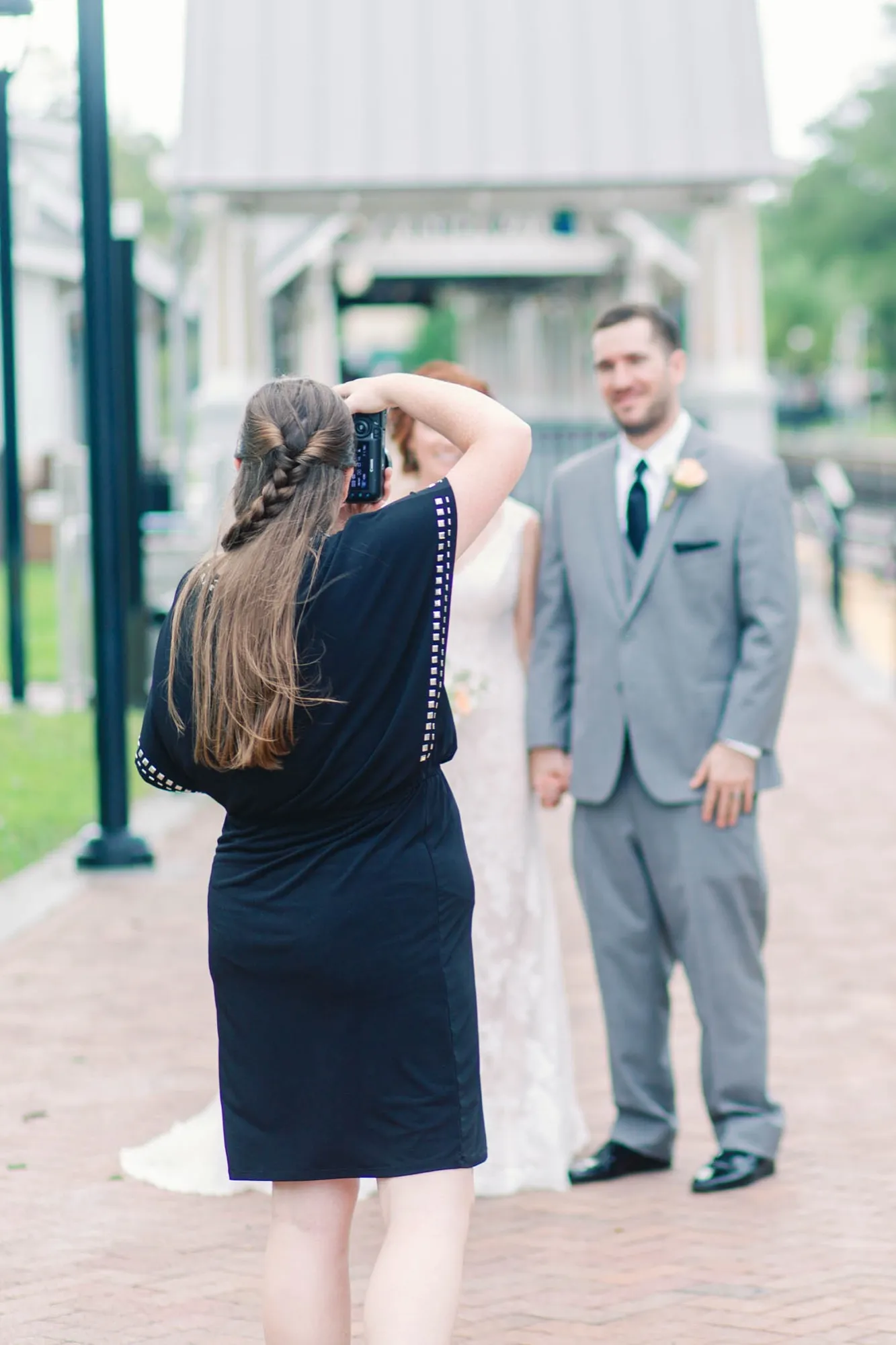 McKenzie in a navy dress raising her camera to photograph a bride and groom on a brick walkway