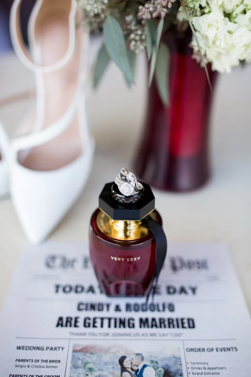 The bride's white heels, a ring box and a newspaper style program laid beside a dark red bouquet
