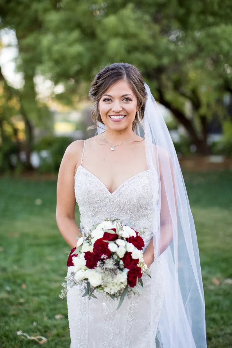 Bridal portrait on the grass with a red and white bouquet and the veil falling behind her